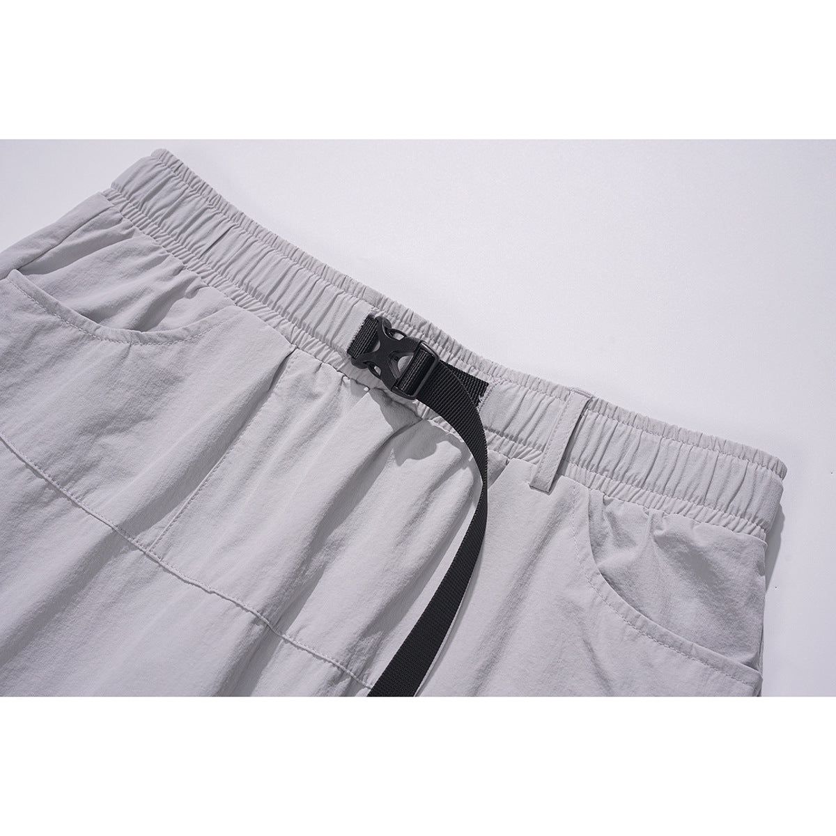 Close-up of light gray shorts showing an elastic waistband, black buckle strap, and curved front pocket.