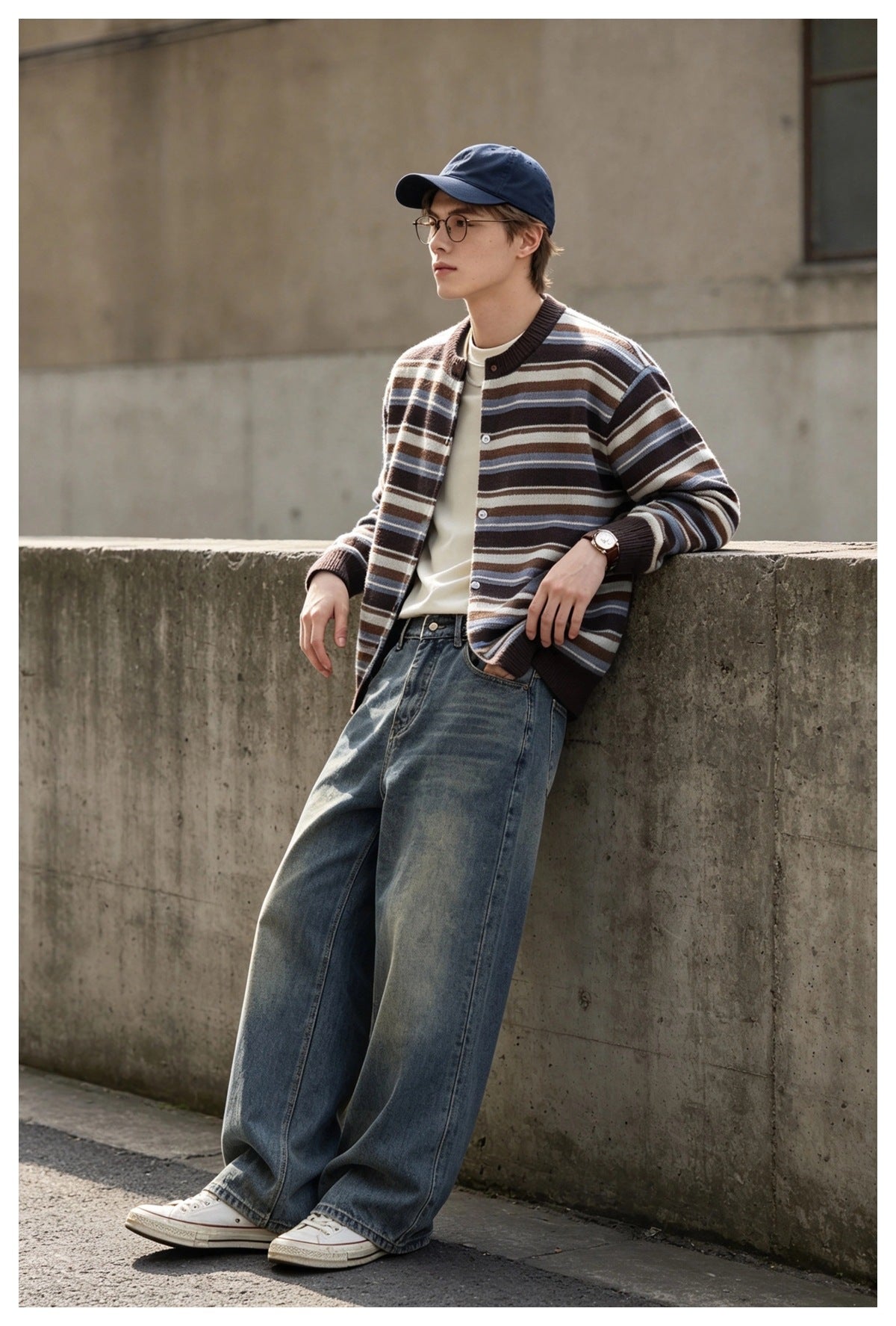 Man leaning on a concrete wall wearing the brown striped cardigan over a cream tee with wide-leg jeans and a navy cap