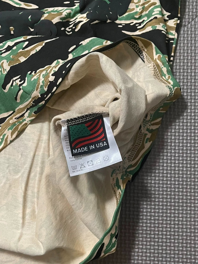 A close-up of the Denim Tears Green Camo All-Over Print T-Shirt shows a tag with a stylized American flag, "MADE IN USA," and care symbols. The shirt is displayed on a textured gray surface.