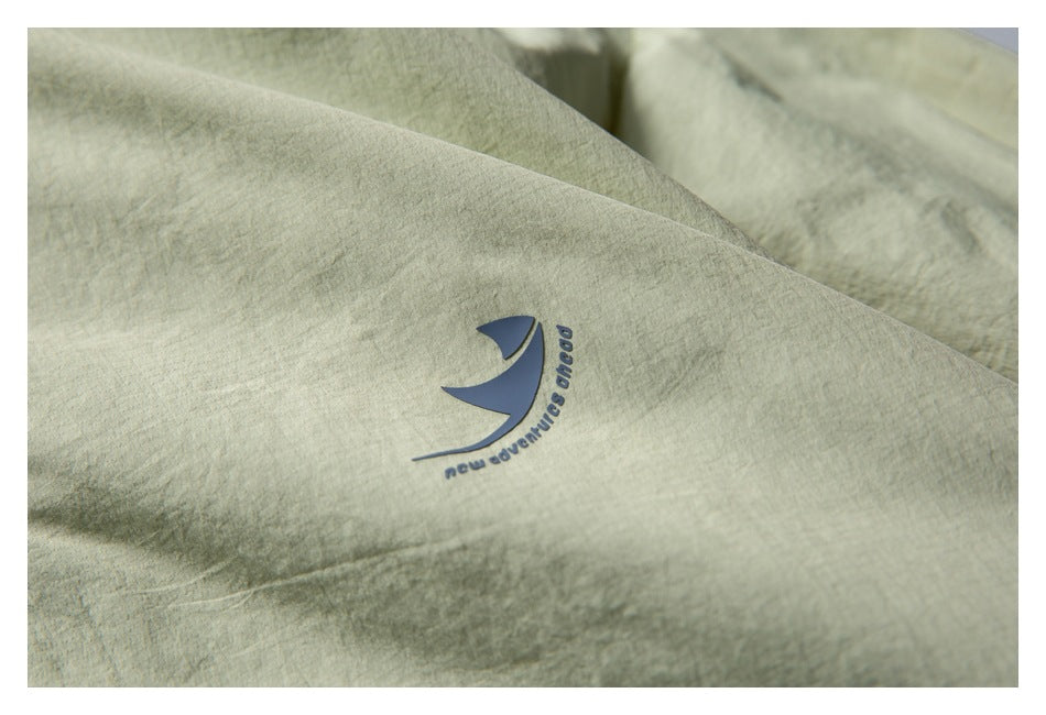 Close-up of a small dark sail-like emblem and English lettering on pale sage jacket fabric.