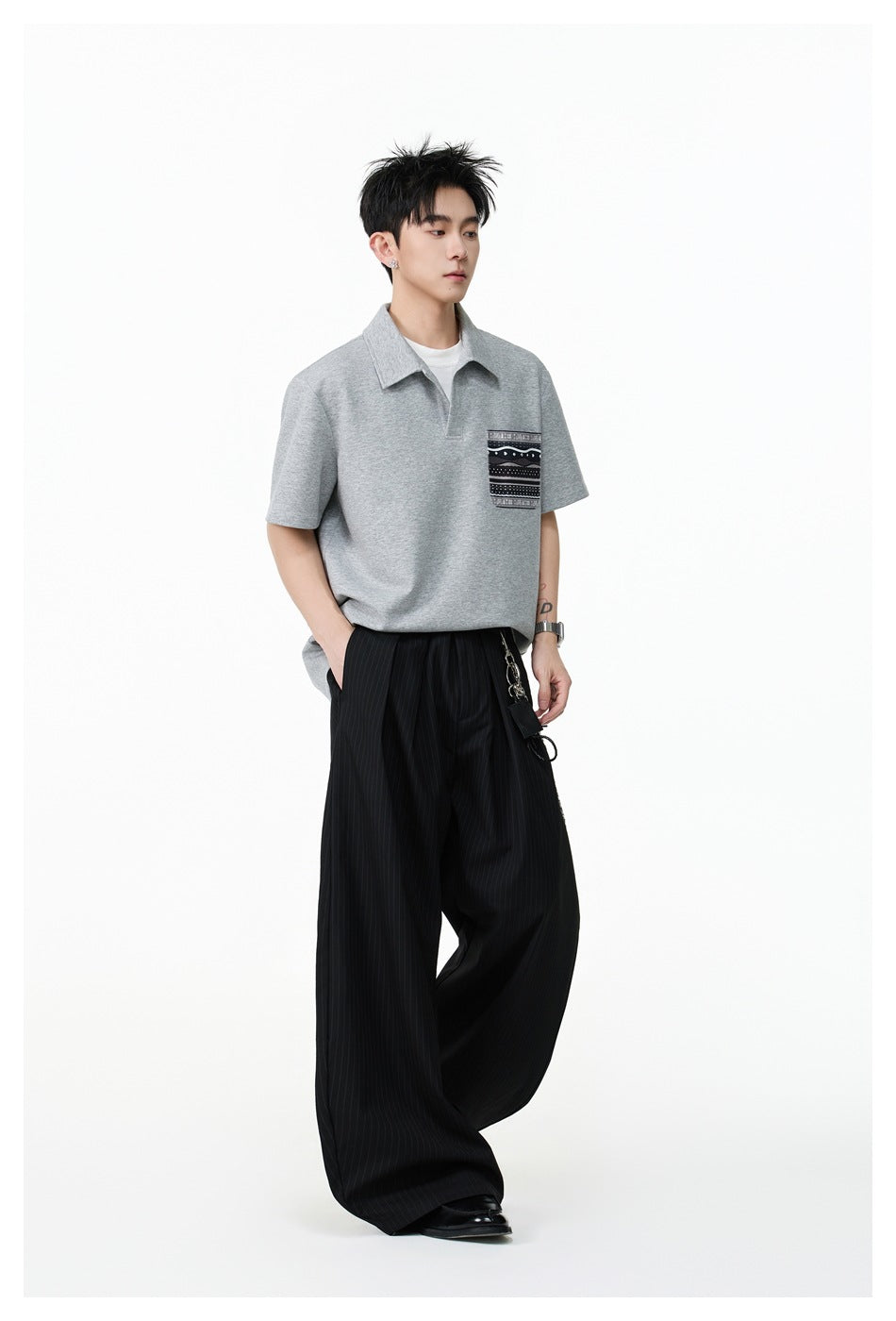 Model wearing a light gray polo with a patterned chest pocket and wide black pinstriped trousers.