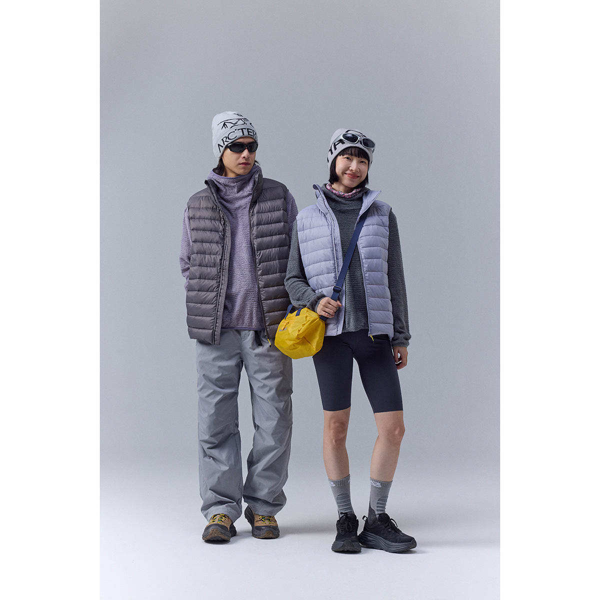 Two models wearing quilted vests: one charcoal gray and one pale lavender, styled with outdoor clothing.