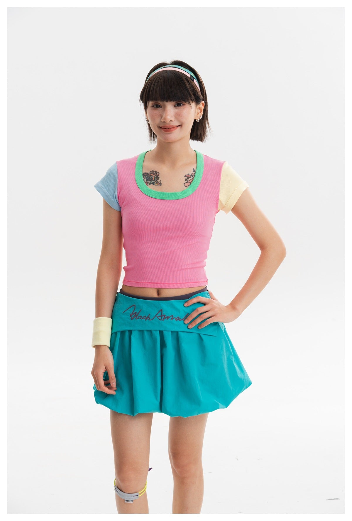 Model wearing a pink cropped top with green trim and mismatched sleeves, paired with a bright teal pleated skirt.