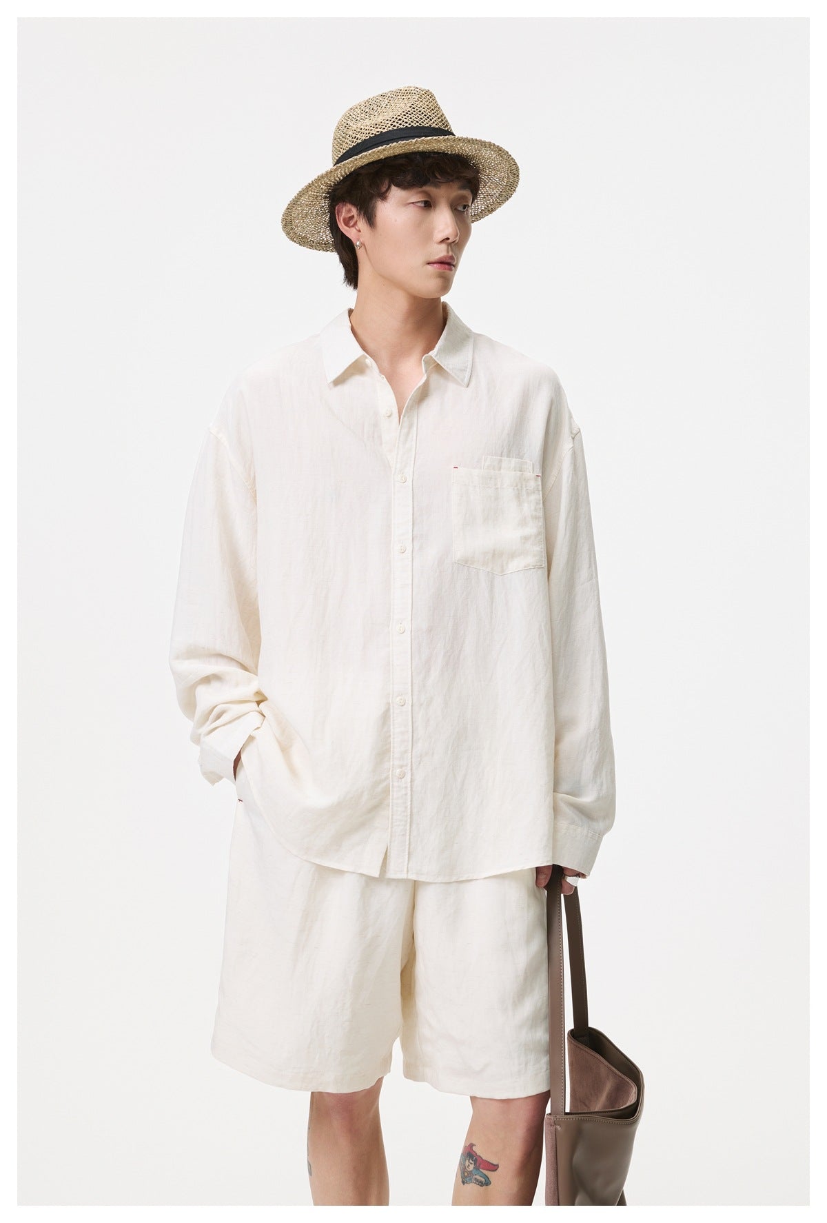 A man models an off-white button-up shirt with a chest pocket and matching shorts, wearing a straw hat.