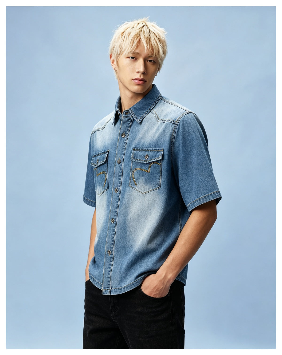Man with blond hair wearing a faded blue denim short-sleeve shirt with black jeans against a light blue background
