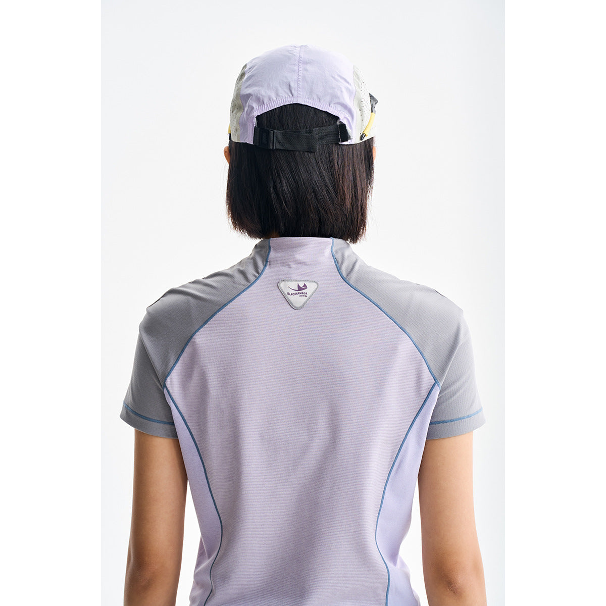 Rear view of a lavender and gray short-sleeve zip-neck top with blue seam piping, worn with a cap.