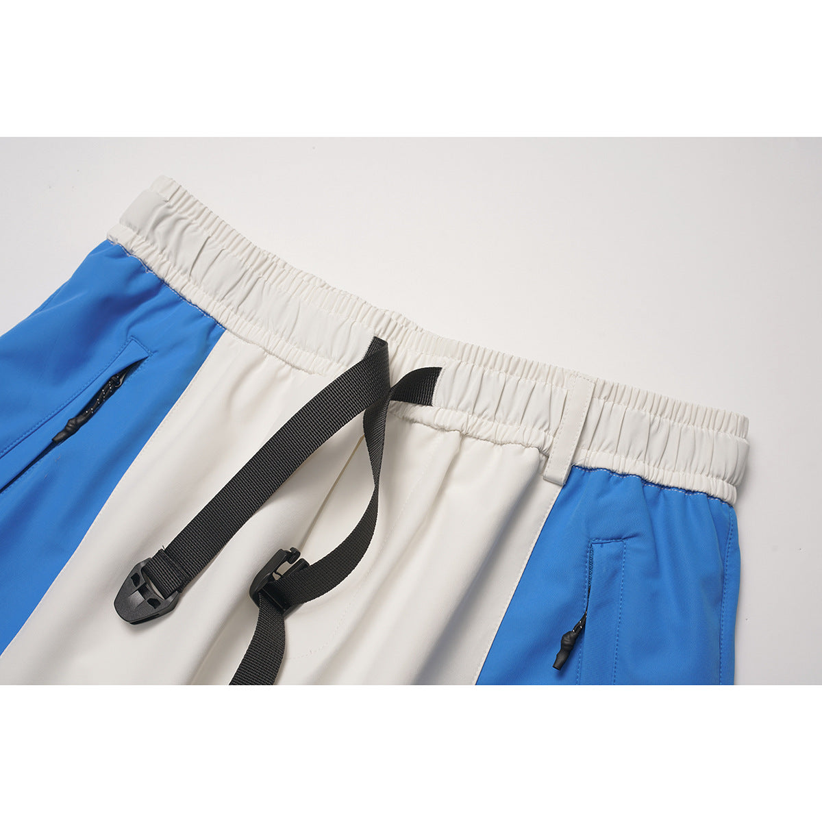 Close-up of blue-and-white pants showing the elastic waistband, black webbing belt and buckle, and zip pockets.