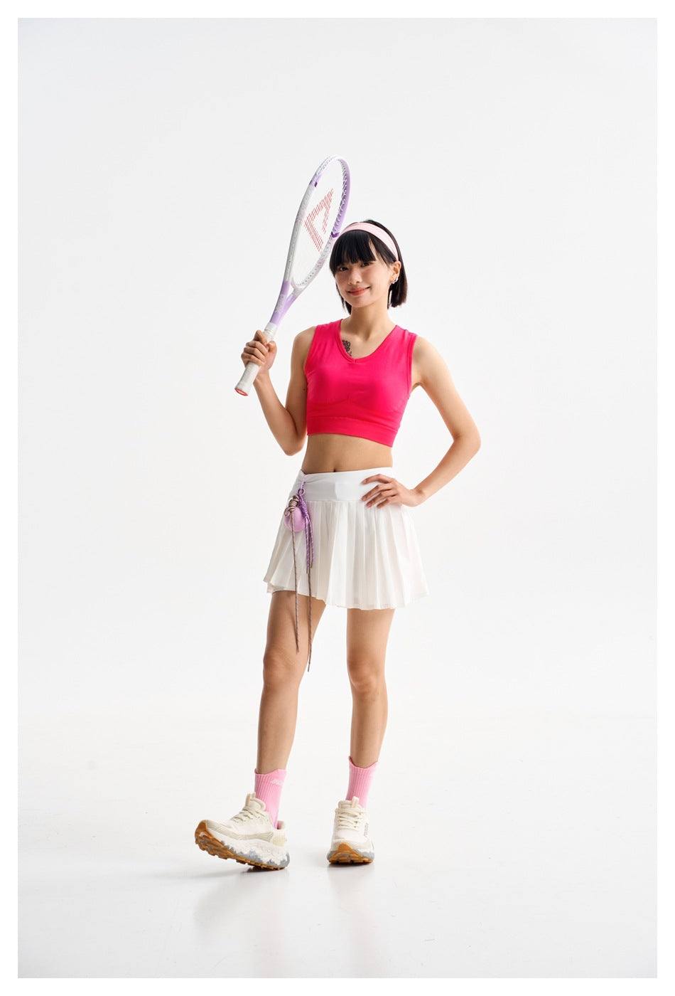 Model wearing a bright pink sleeveless crop top and pleated white tennis skirt, holding a racket.