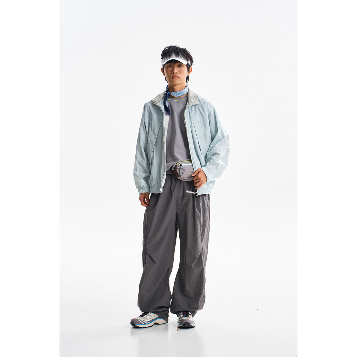 A model wears a pale blue zip jacket with dark gray loose trousers, a white visor, and a blue neck scarf.