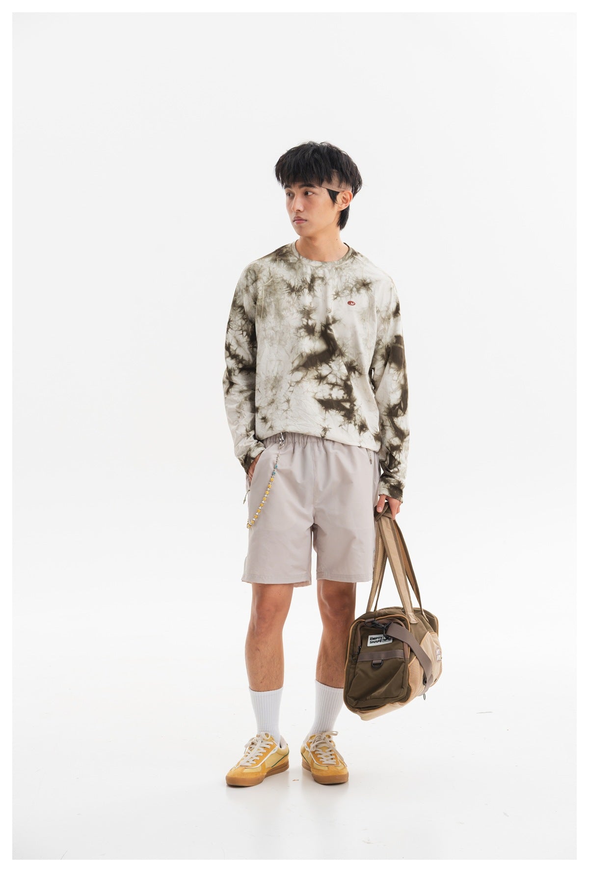 Model wearing an olive-gray tie-dye long-sleeve shirt and pale gray shorts, holding a brown duffel bag.
