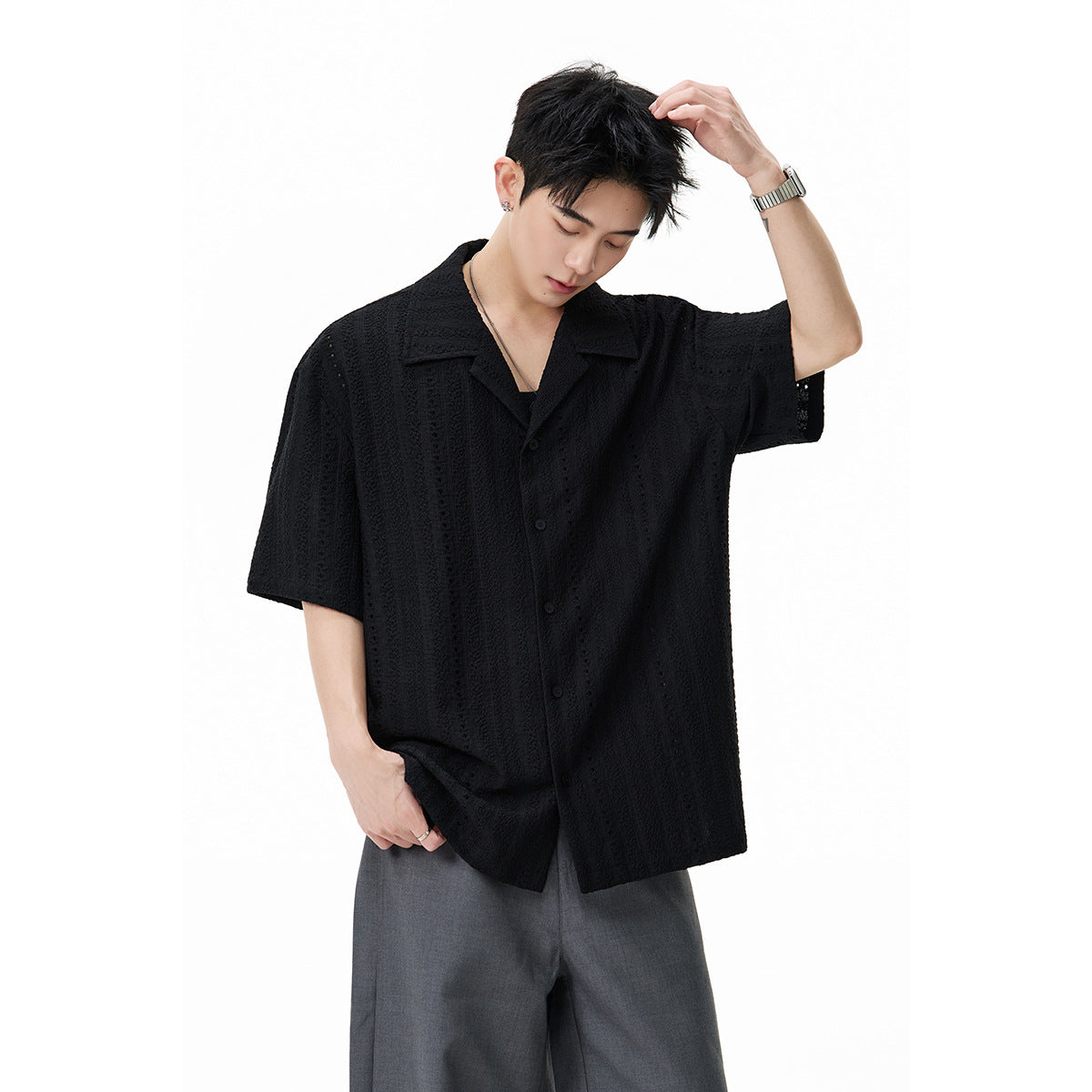 Model wearing a black short-sleeve eyelet shirt with a notched collar and gray trousers against a white background.