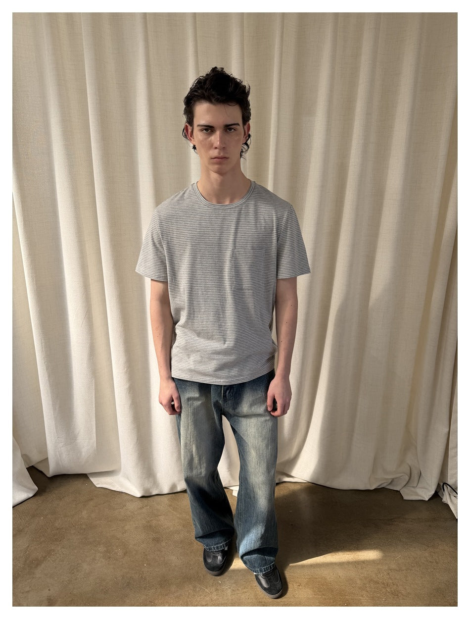 Model wearing gray striped short-sleeve pocket T-shirt with wide-leg blue jeans, standing in front of a cream curtain