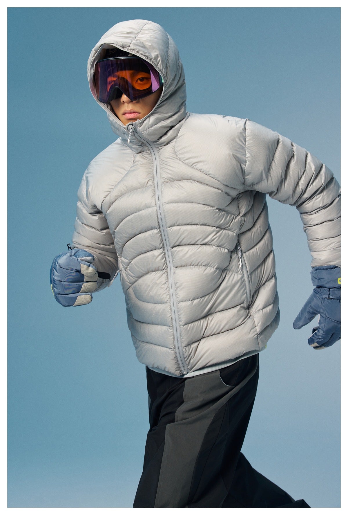 Model wearing a light gray quilted puffer jacket with the hood up and blue ski gloves.