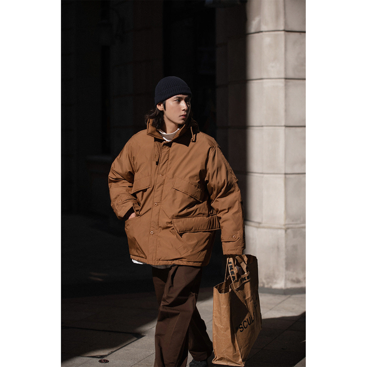 Model wearing a brown hooded jacket with button closure and multiple flap pockets, styled with dark trousers and a navy