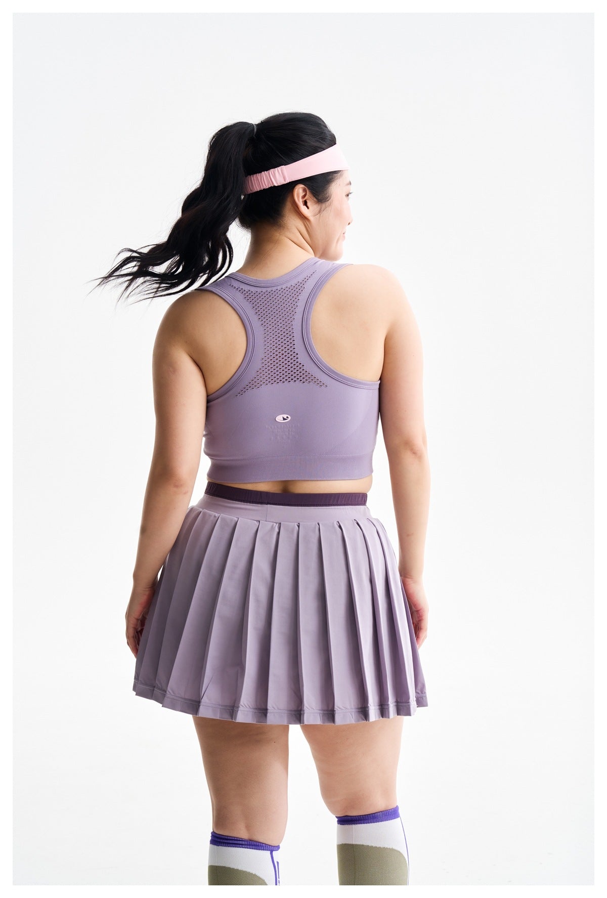 Back view of a model wearing a lavender racerback athletic top with mesh detailing and a pleated skirt.