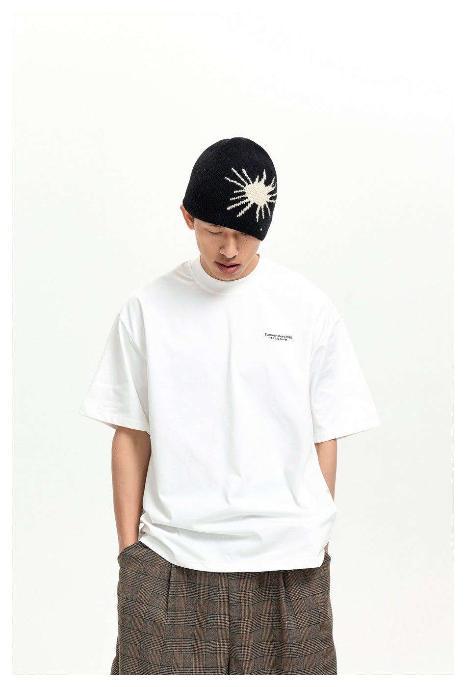 Model wearing a white boxy T-shirt with small chest text, a black sun-pattern beanie and plaid trousers, close view