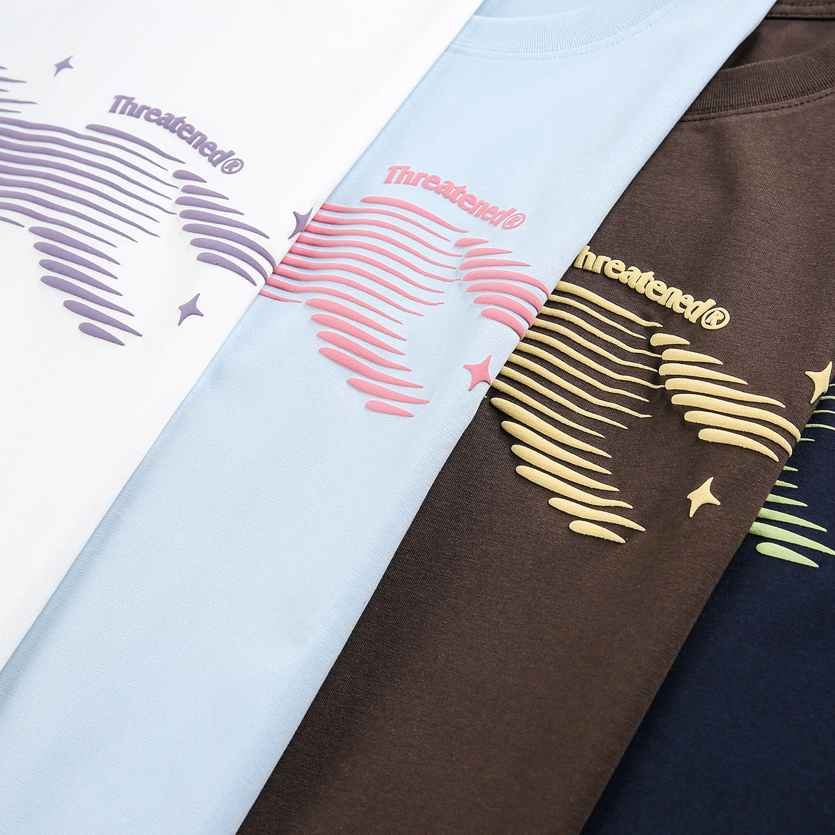 Close-up of four graphic T-shirts in white, pale blue, brown, and navy, with contrasting raised prints.