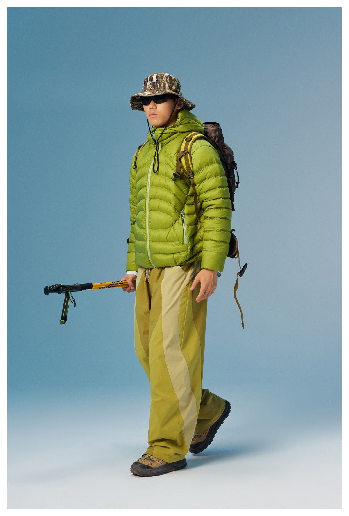 Full-length model wearing a lime-green quilted hooded jacket with a backpack and hiking gear.
