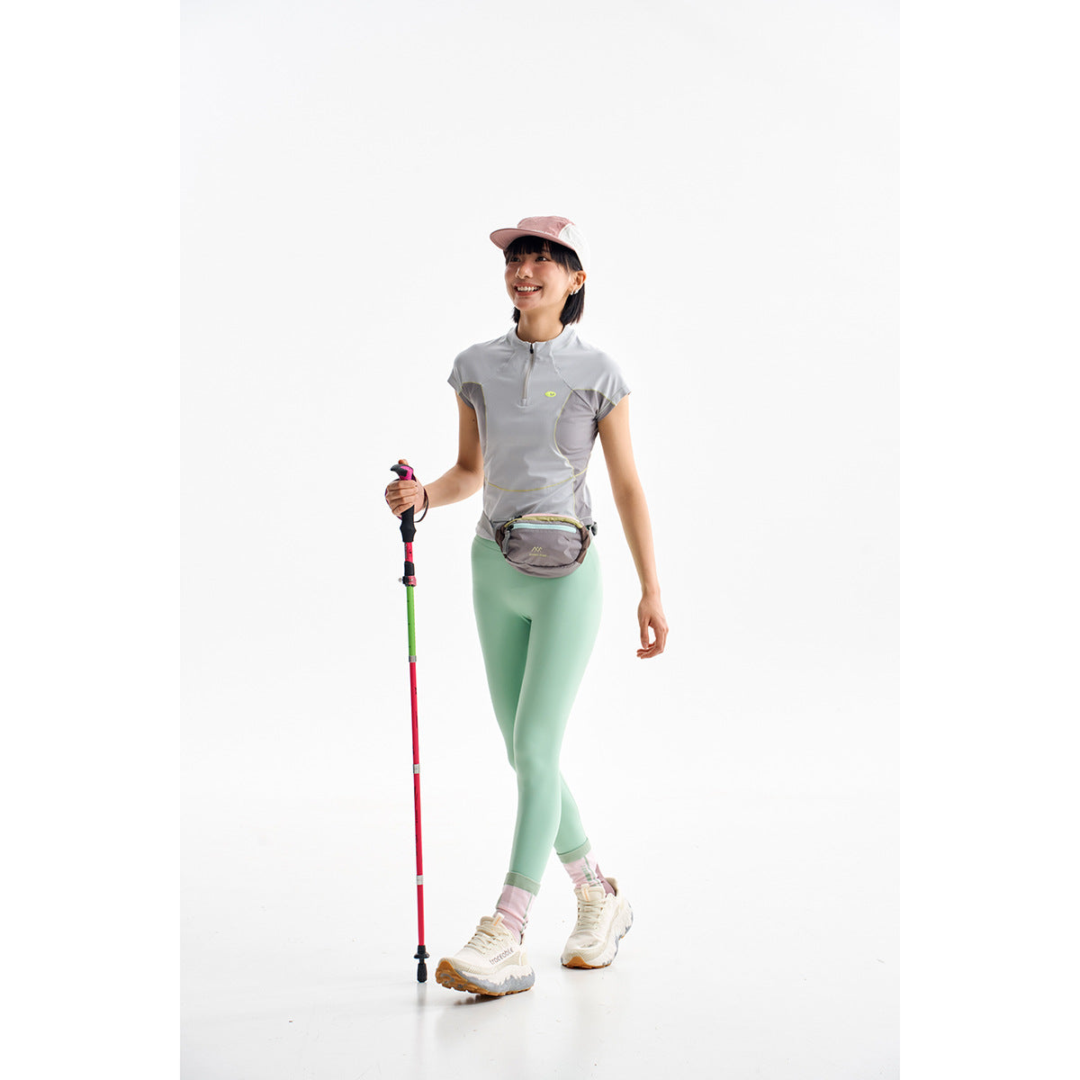 Woman in a gray short-sleeve zip top and mint leggings, holding a trekking pole against a white background.