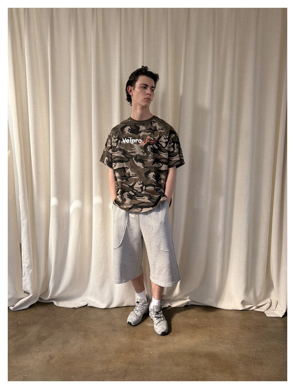 Man wearing light khaki camouflage T-shirt with gray shorts and sneakers, standing in front of a cream curtain