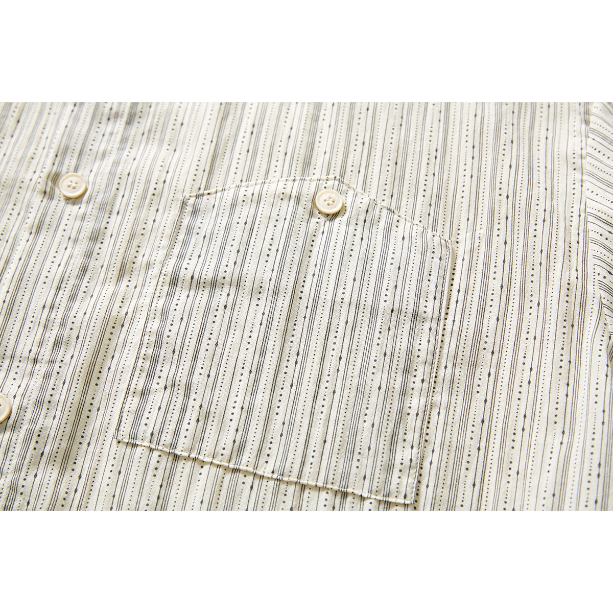 Close-up of the shirt’s single chest pocket and button on ivory fabric with fine black dotted stripes.