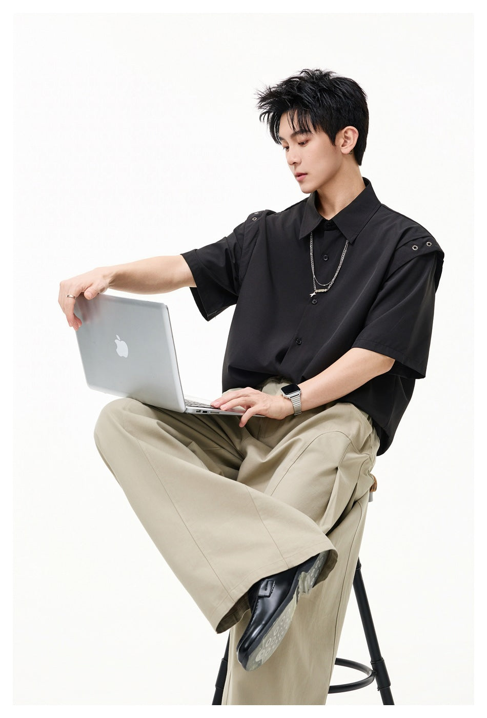 Seated model wearing a black short-sleeve button-up shirt with metal-eyelet shoulder tabs and beige wide-leg trousers.