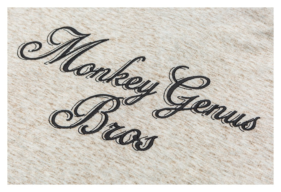 Close-up of black cursive “Monkey Genius Bros” embroidery on beige marled fabric.