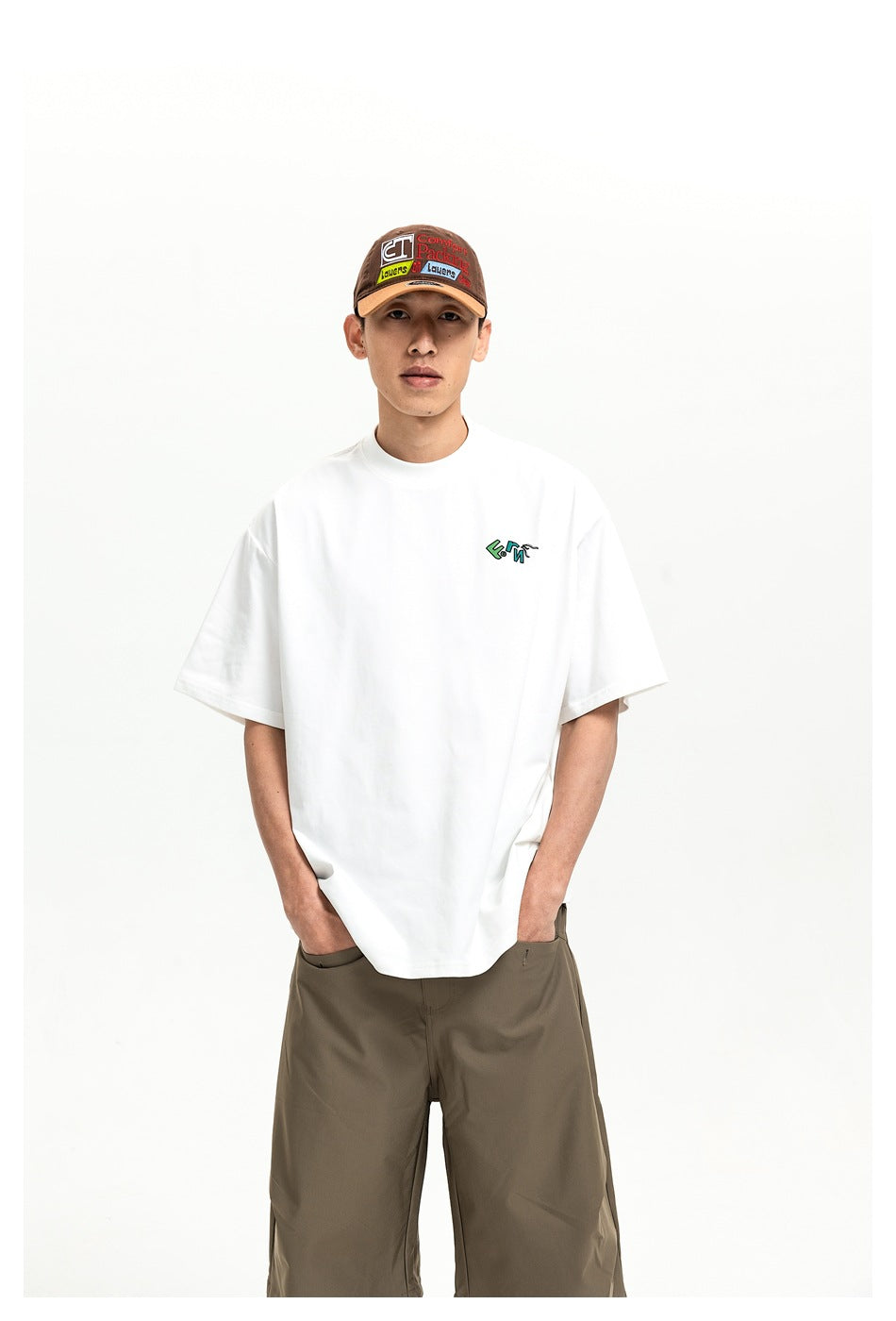 Person wearing a white oversized crew-neck T-shirt with a small green chest logo, olive pants and a brown cap