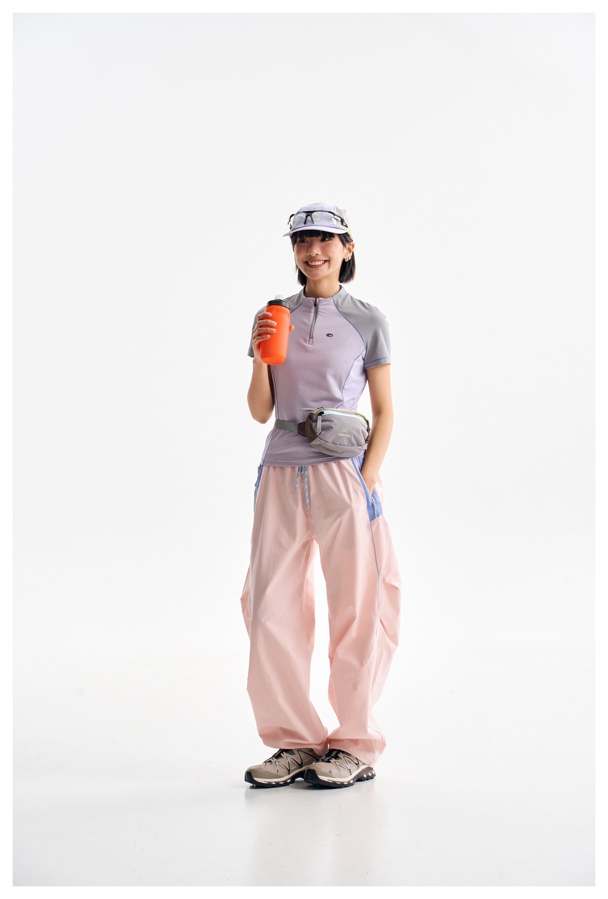 Model wearing a lavender and gray zip-neck top with loose pink trousers, a waist pack, cap, and trail shoes.