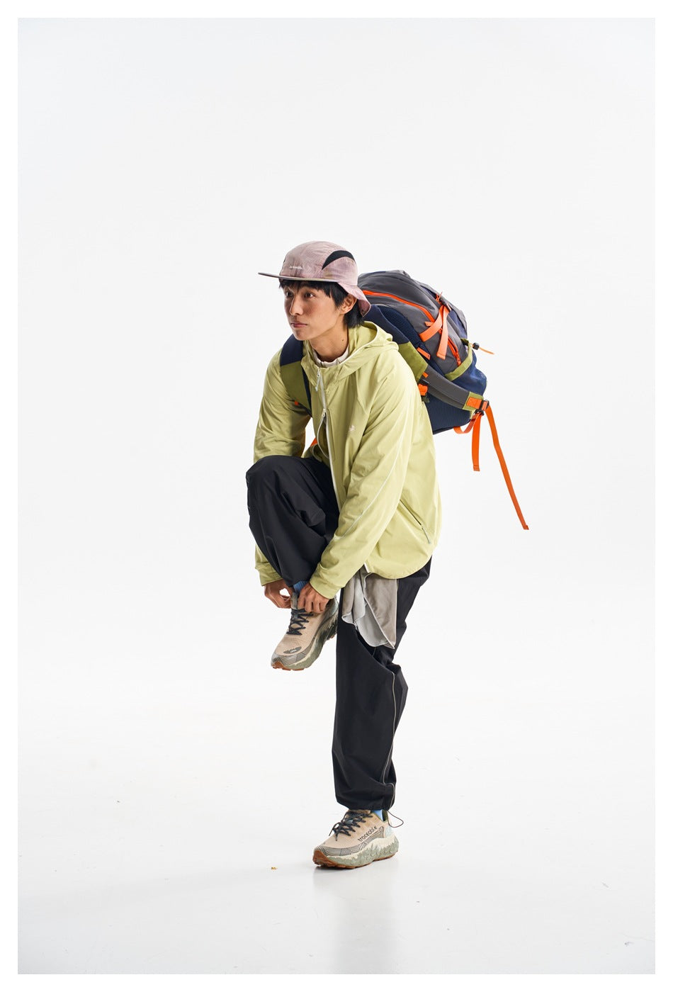 Model crouching in a pale yellow-green hooded zip jacket and black trousers with a hiking backpack.