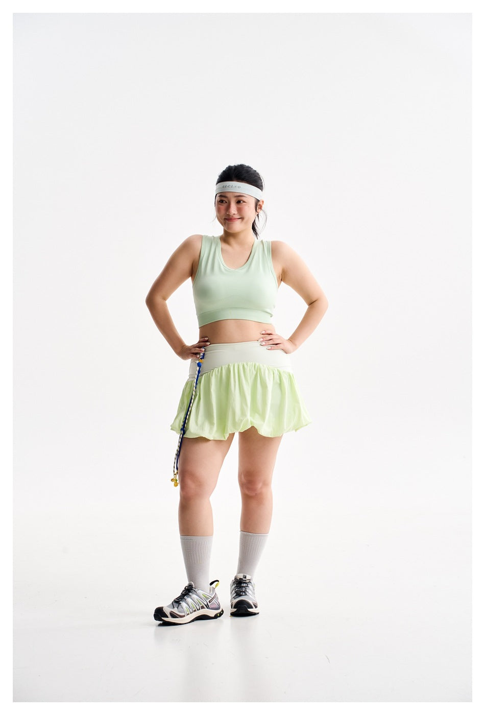 Model wearing a mint-green cropped sports bra and pale lime flared skirt with a light headband and gray socks.