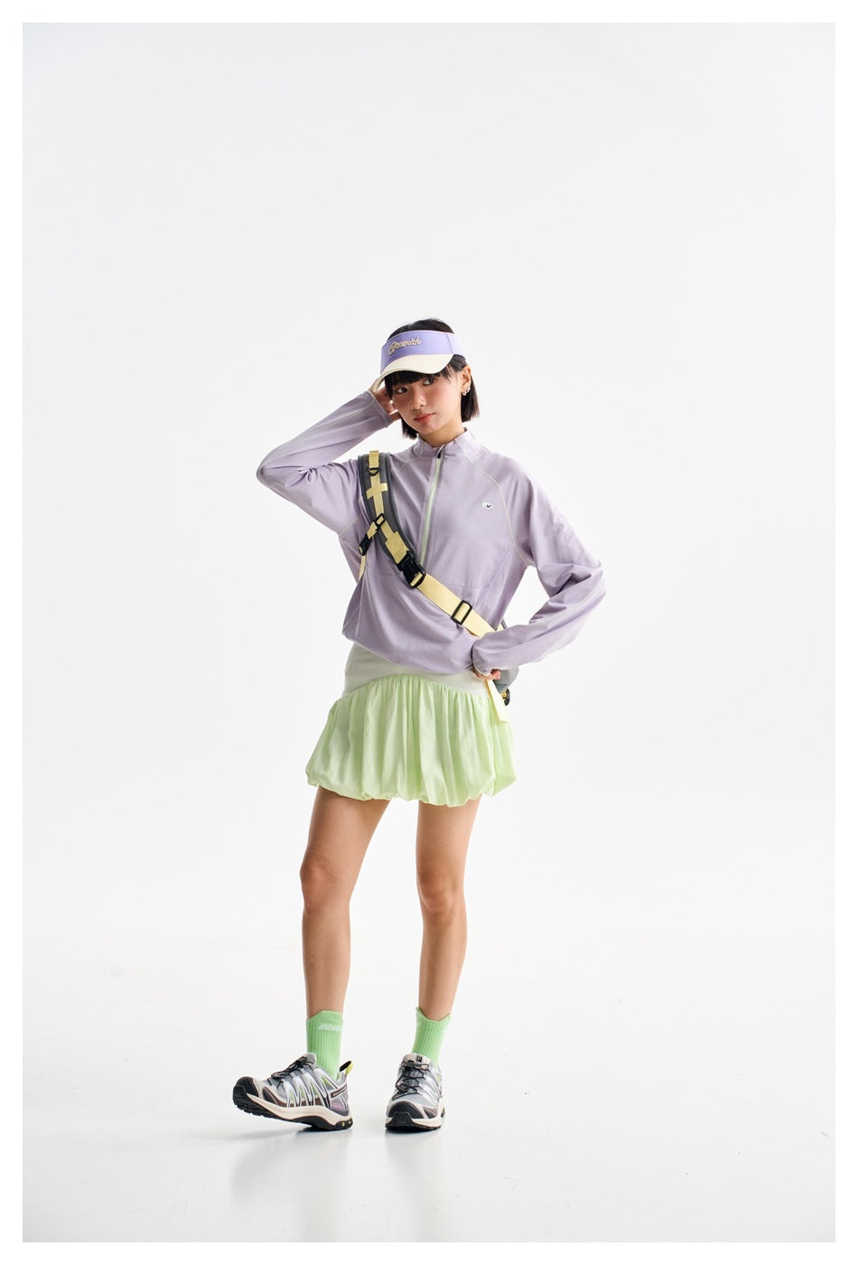 Woman posing in a lavender quarter-zip top and pale green skirt with a crossbody strap.