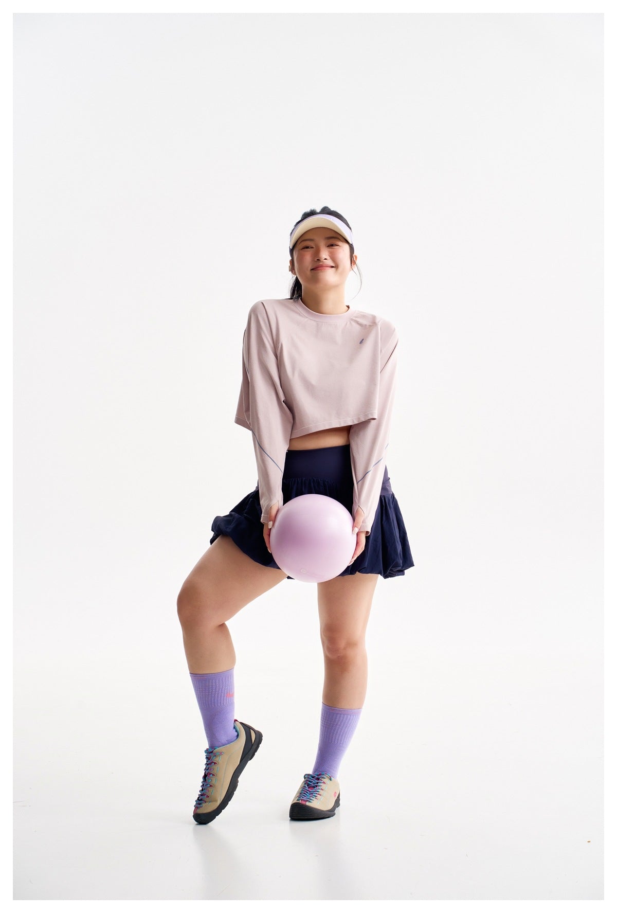 Model wearing a pale pink cropped long-sleeve top and navy bubble-hem skirt, with lavender socks and a visor.