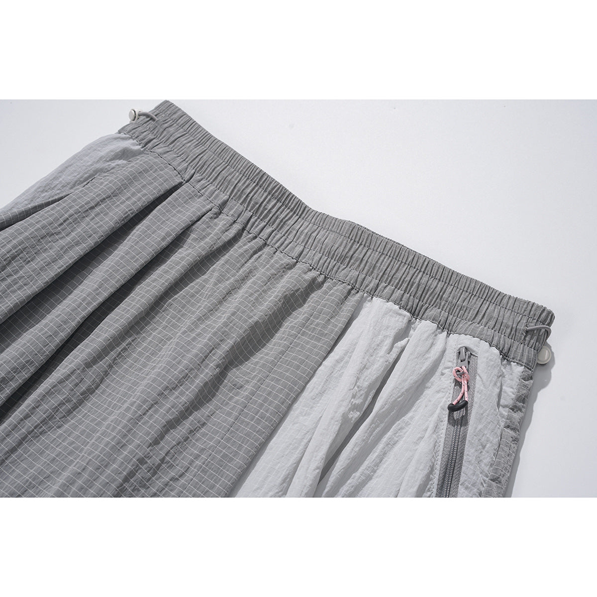 Close-up of gray pants showing the gathered elastic waistband, grid-patterned panels, and a zip pocket with a pink pull.