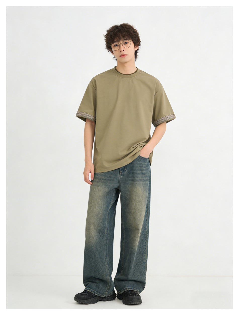 Model wearing an olive-khaki short-sleeve T-shirt with plaid cuff trim and loose blue jeans, shown full length.