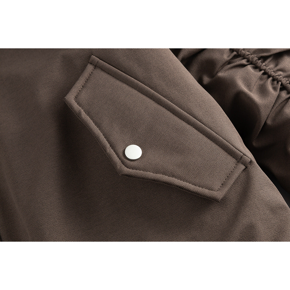 Close-up of a brown jacket's flap pocket with a white snap button and gathered sleeve detail.