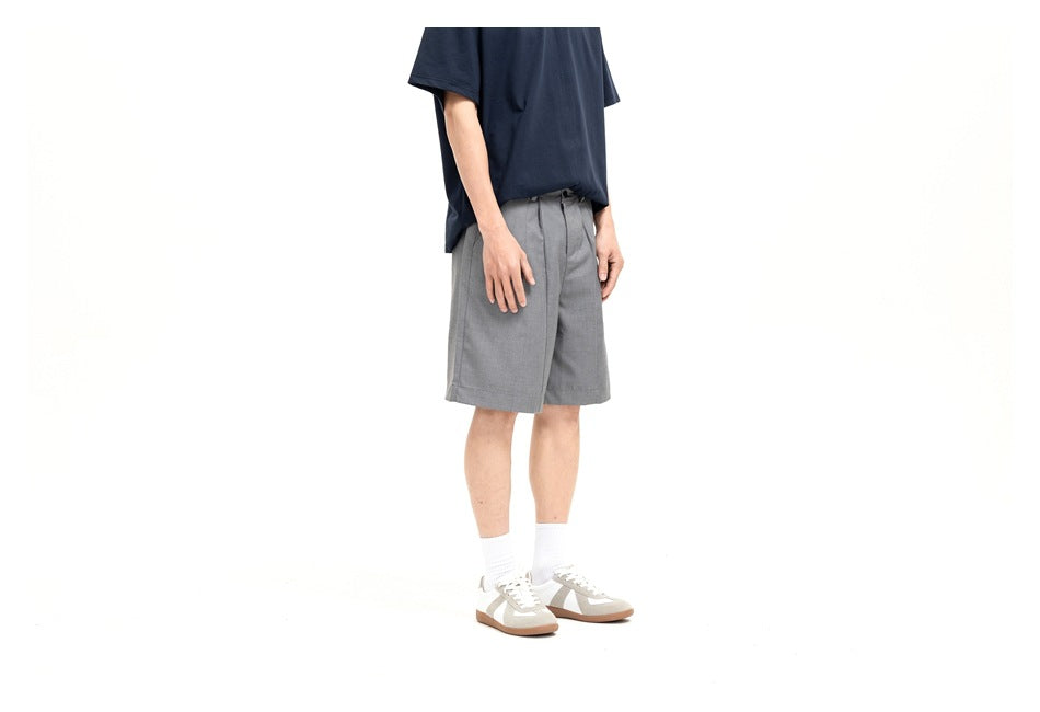 Man wearing gray knee-length pleated shorts with a navy t-shirt and white sneakers, side view