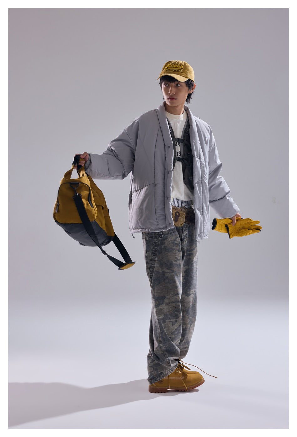 Model wearing the gray padded jacket with camouflage pants, holding a yellow bag and gloves