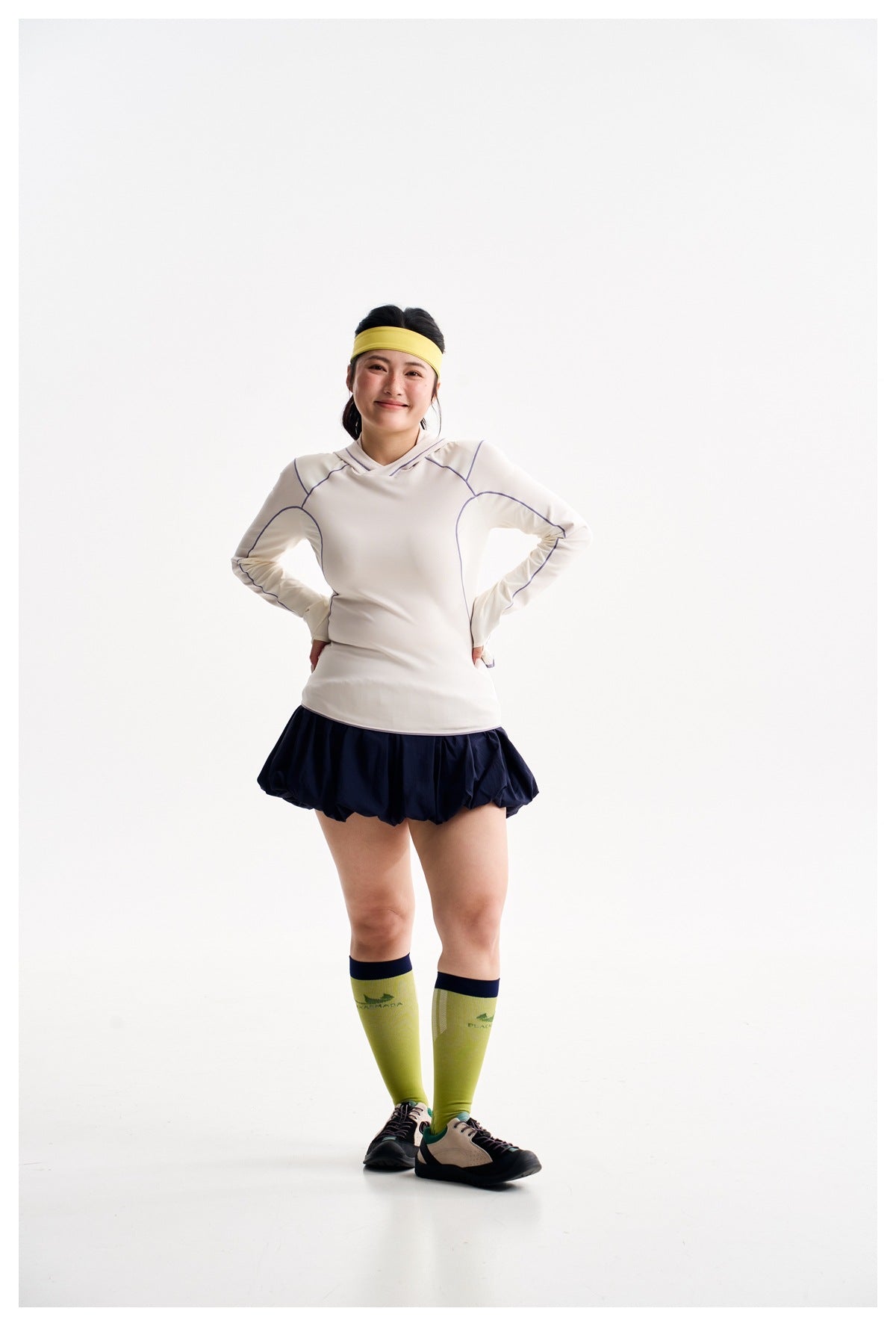 Full-length model wearing an ivory hooded top with purple piping, navy bubble skirt, lime knee socks and yellow headband.