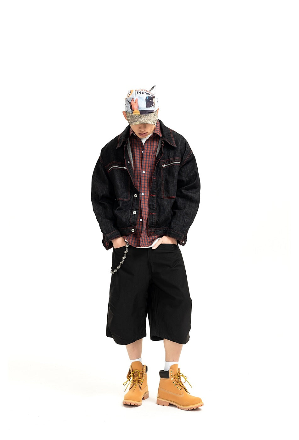 Man in a black denim jacket over a plaid shirt with black wide shorts, printed cap and tan boots, full-length view