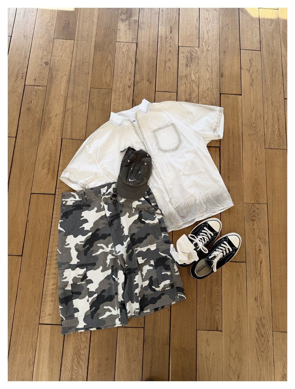 White short-sleeve shirt with chest pocket laid flat on wood floor with camo shorts, cap and sneakers