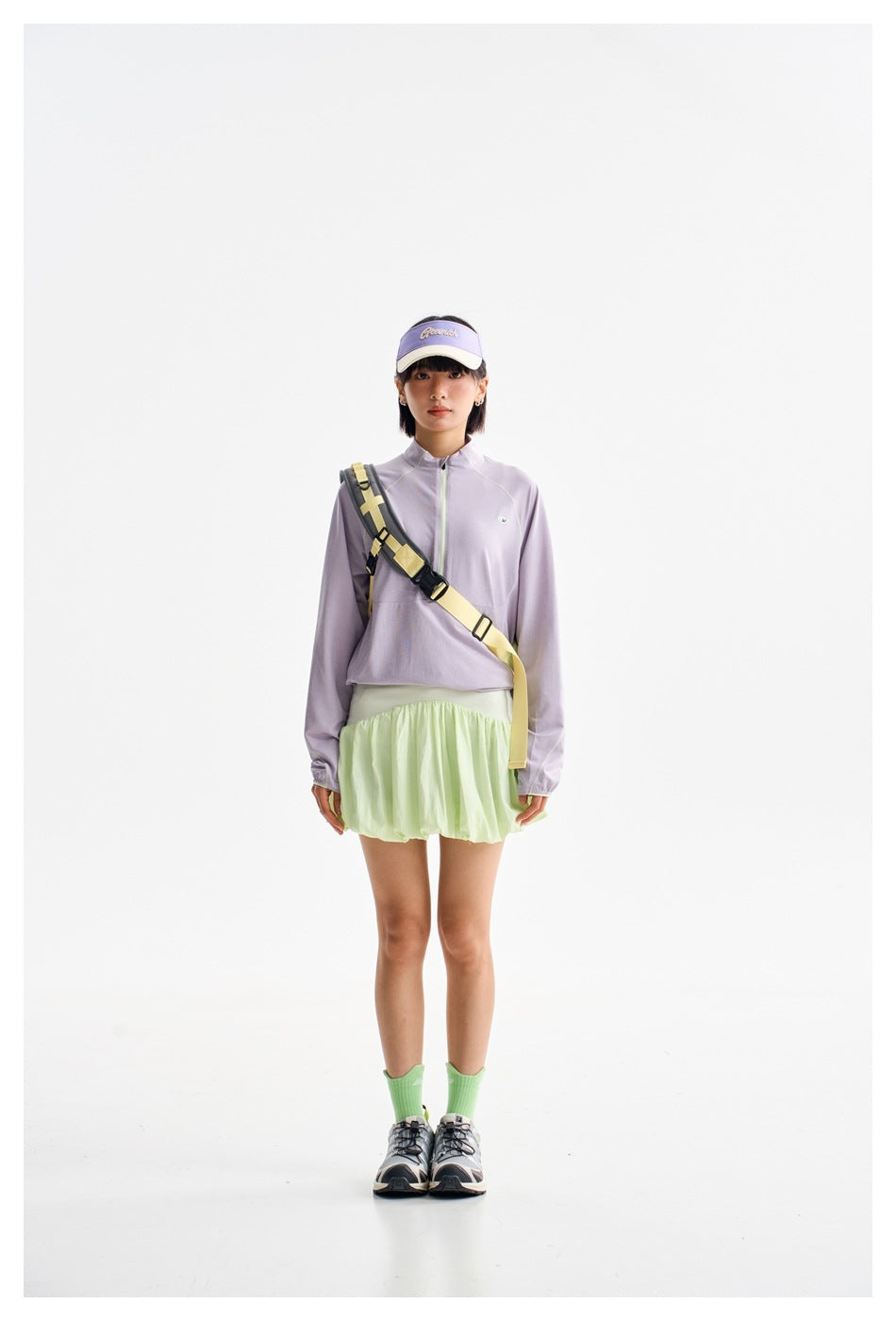 Woman facing forward in a lavender quarter-zip top and pale green skirt with a crossbody strap.