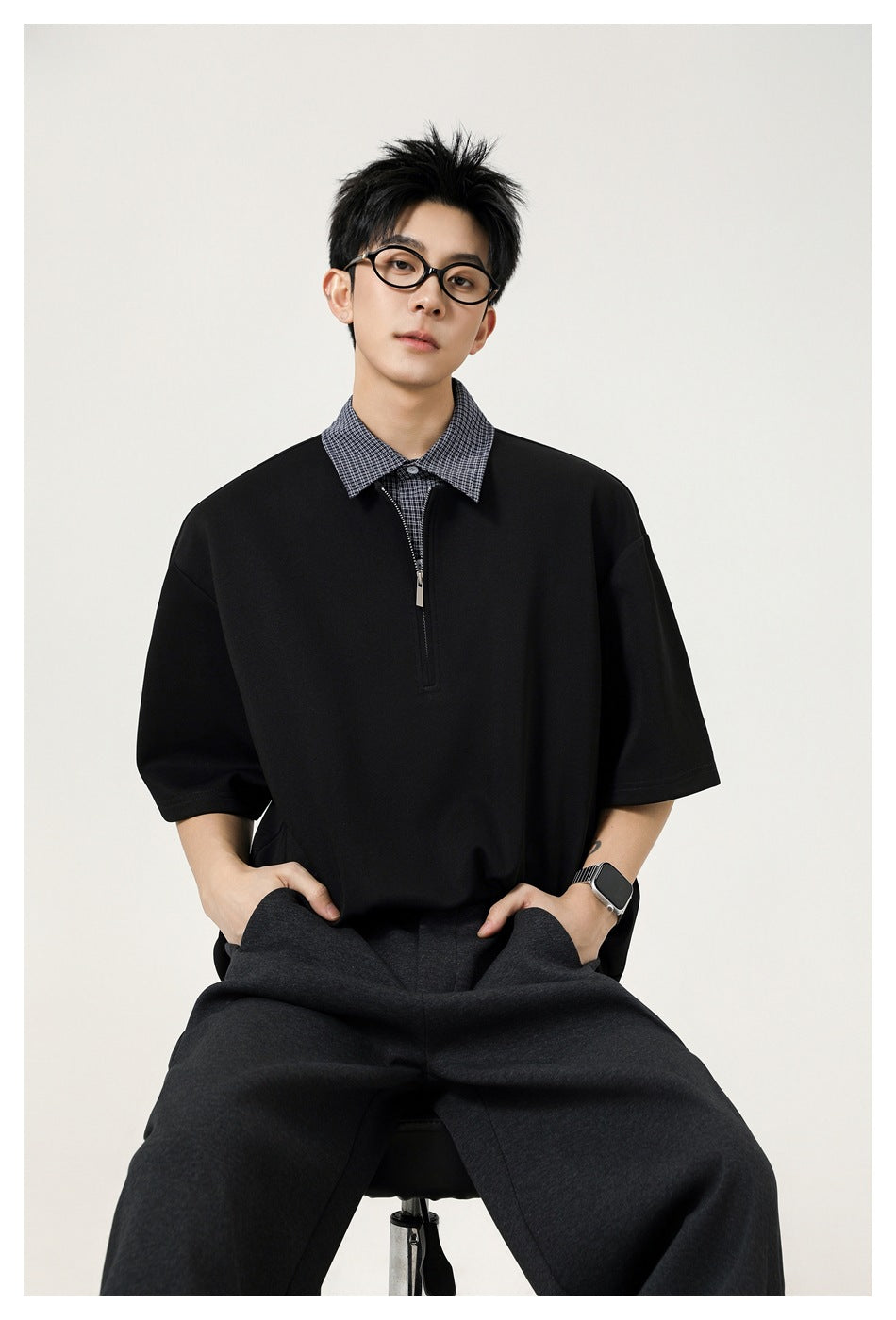 Seated male model wearing a black oversized short-sleeve top with a checked collar and short zip placket.