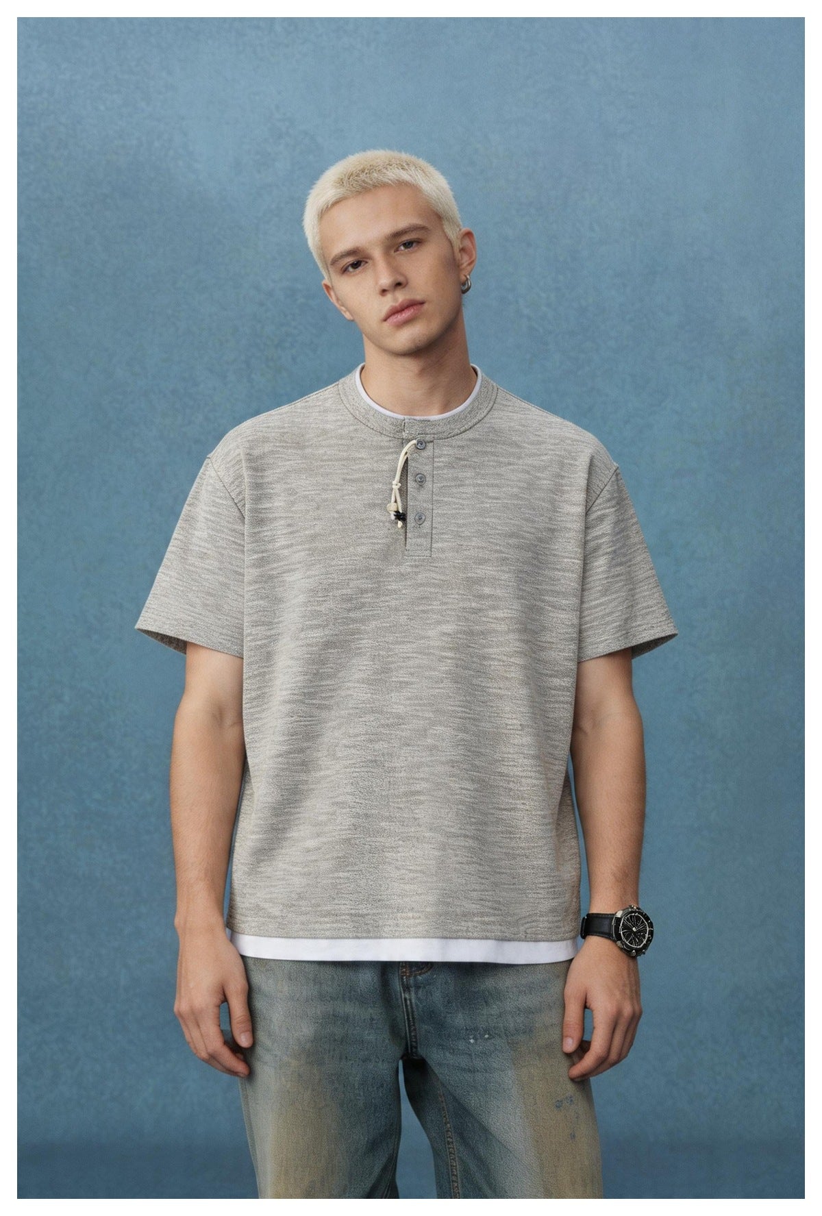 Man wearing a heathered gray short-sleeve henley T-shirt with beaded cord and white layer at hem, with faded jeans