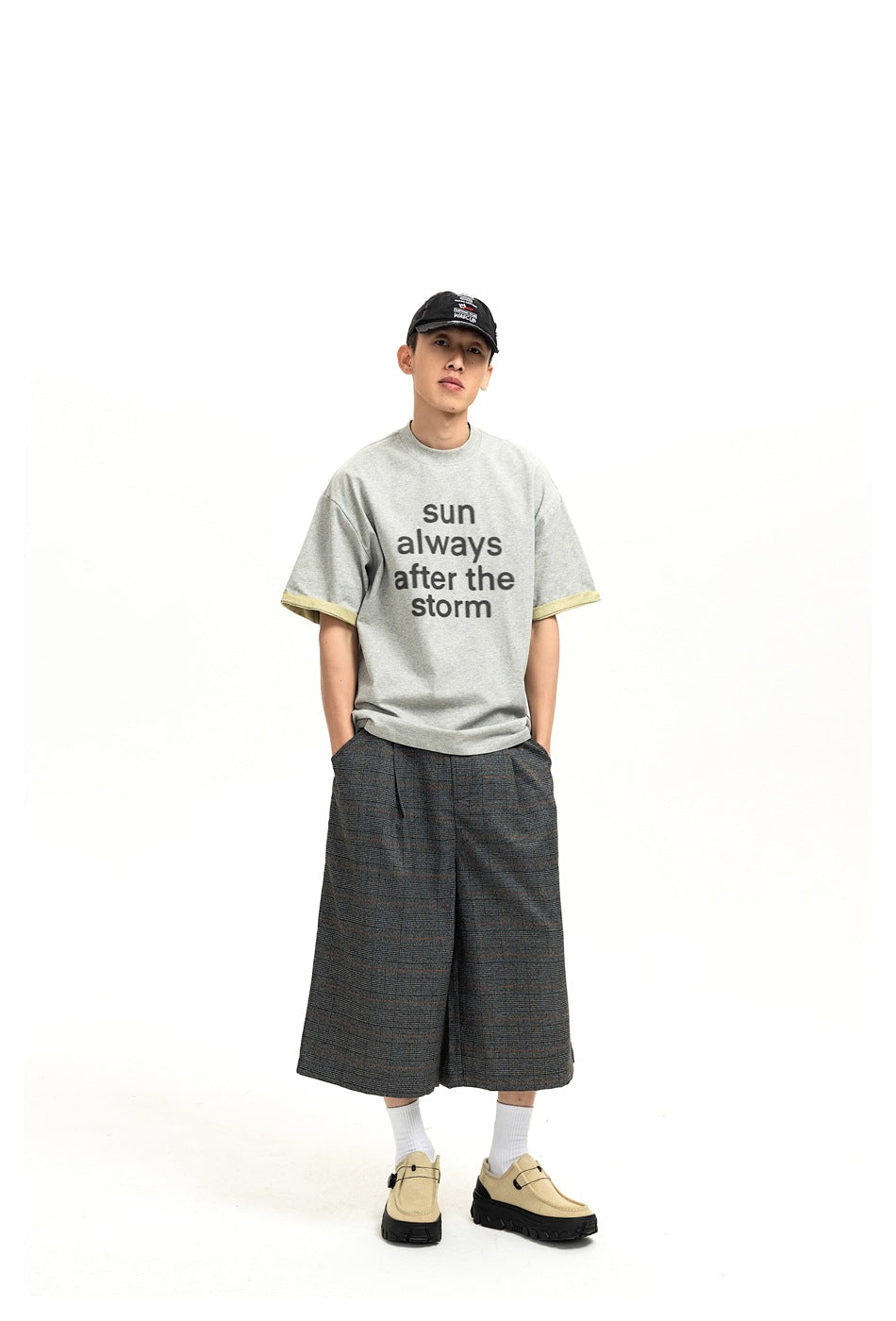 Model wearing gray 'sun always after the storm' T-shirt with yellow cuffs, plaid wide-leg shorts, cap and beige shoes