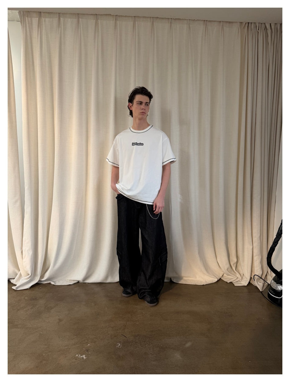 Full-length model wearing a white T-shirt with black contrast stitching and loose black jeans.