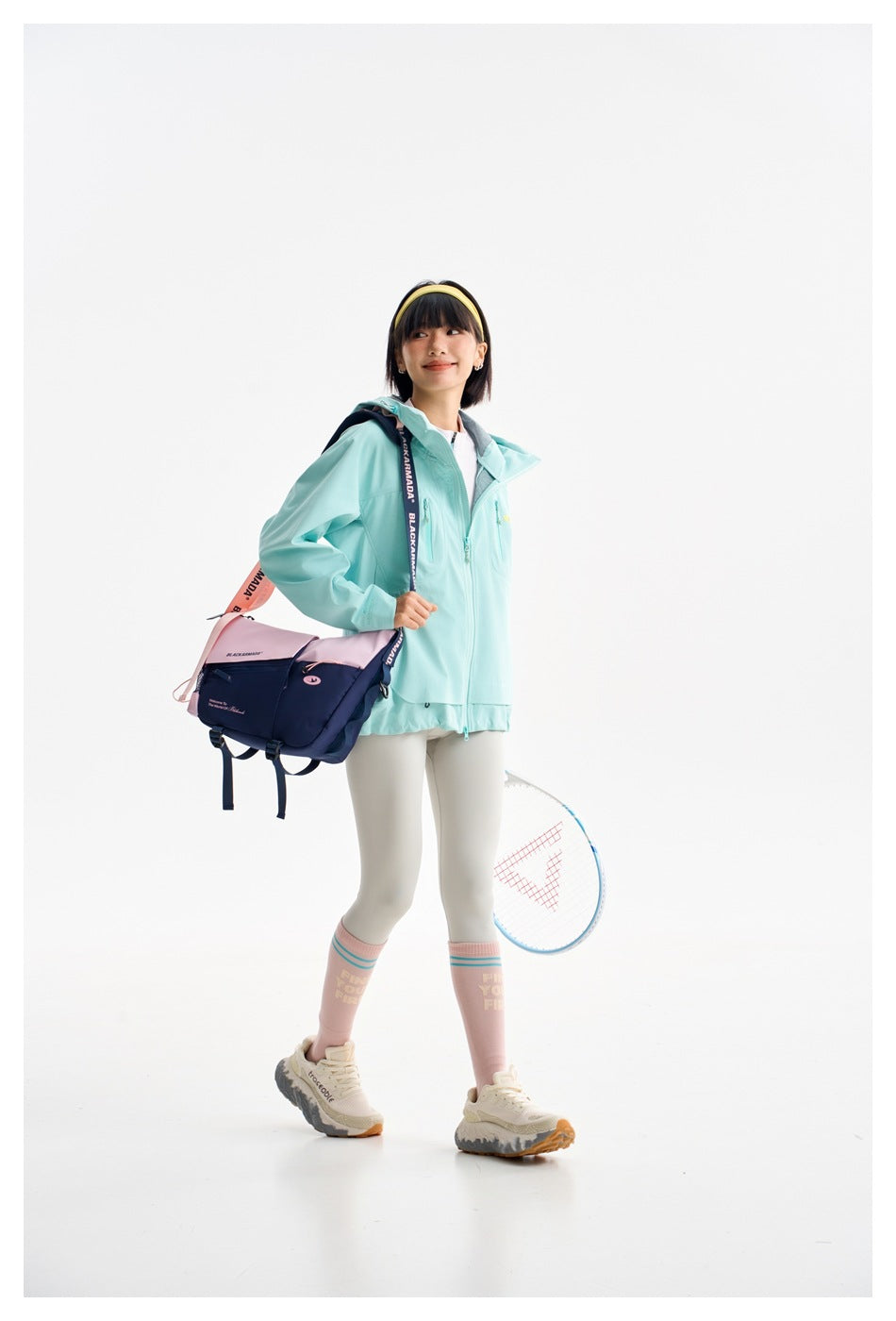Model in a mint hooded jacket and white leggings, carrying a navy and pink shoulder bag with a tennis racket.