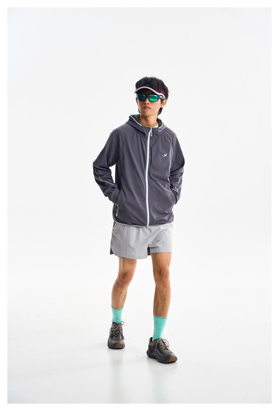 Model facing forward in a charcoal hooded zip jacket with light trim and light gray shorts.