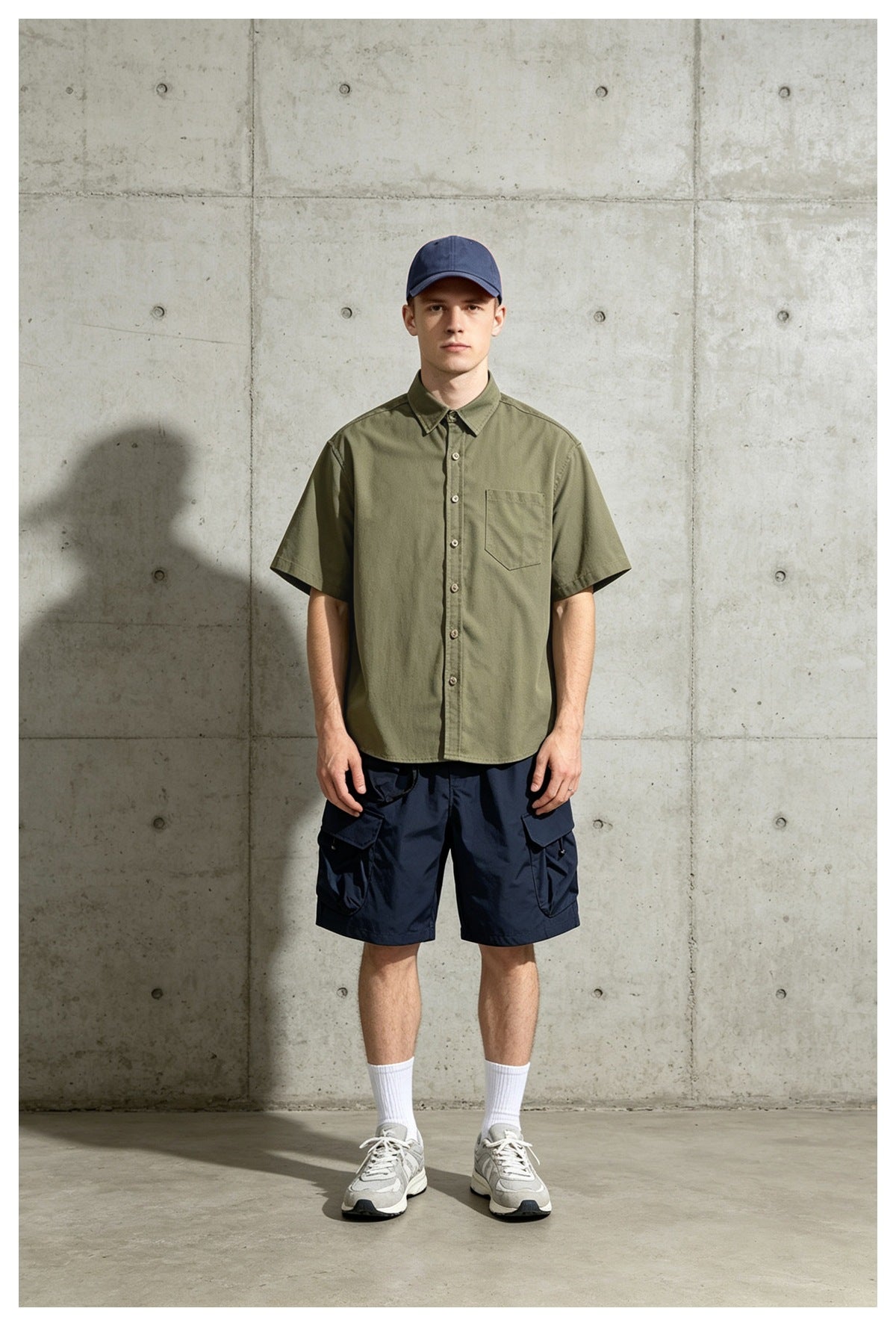 Man in army green short-sleeve shirt, navy cap and navy cargo shorts standing against a concrete wall