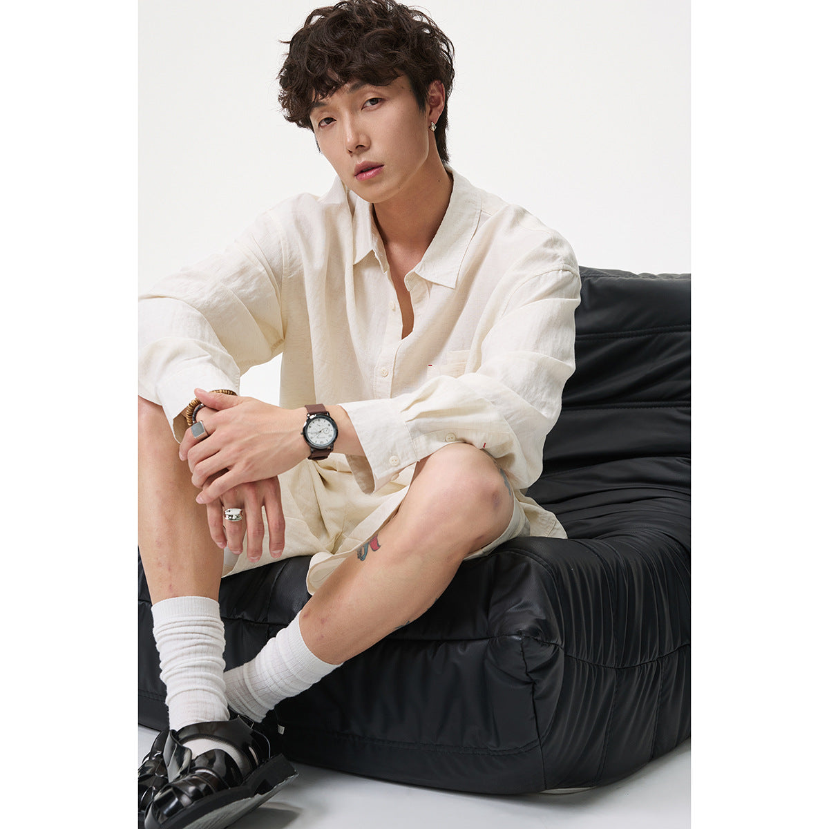A seated man wears an off-white long-sleeve button-up shirt with a chest pocket and matching shorts.